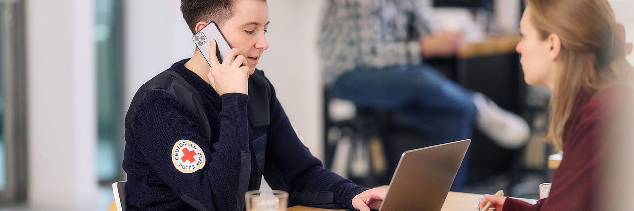DRK-Mitarbeiterin telefoniert mit Handy und schaut auf Laptop.